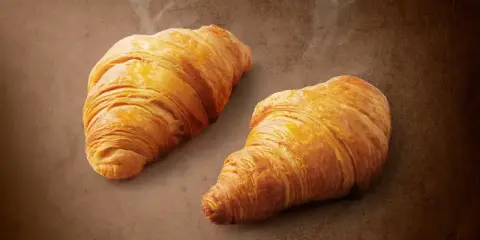 Dwa złociste, parujące croissanty na brązowym tle. Pyszne, choć warto pamiętać o ich kaloryczności.