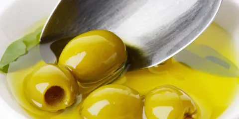 Kilka zielonych oliwek w złocistej oliwie, łyżka zanurzona w płynie. Oliwa z oliwek kcal - zdrowie i smak.