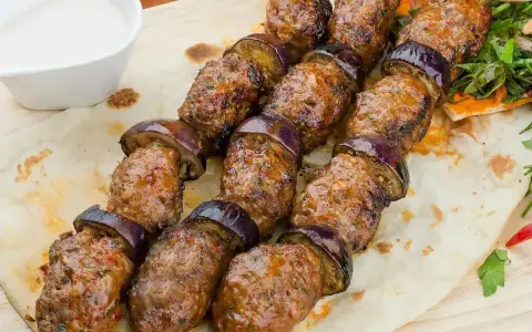 Kofta kebab z bakłażanem na patyku, podany na płaskim chlebie z sosem.