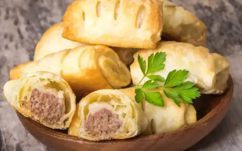 Stos pieczonego ciasta francuskiego z kiełbaską w środku, ozdobiony gałązką pietruszki.