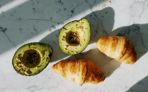 Dwa połówki awokado z nasionami i dwa chrupiące croissanty na marmurowym blacie. Idealne składniki na pyszne awokado przepisy.
