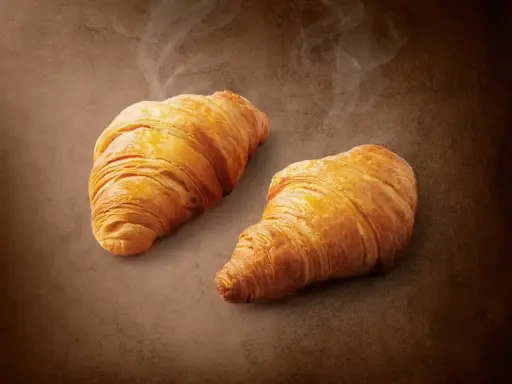 Dwa złociste, parujące croissanty na brązowym tle. Pyszne, choć warto pamiętać o ich kaloryczności.