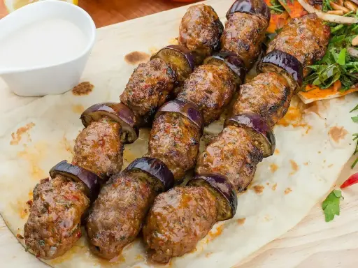Kofta kebab z bakłażanem na patyku, podany na płaskim chlebie z sosem.