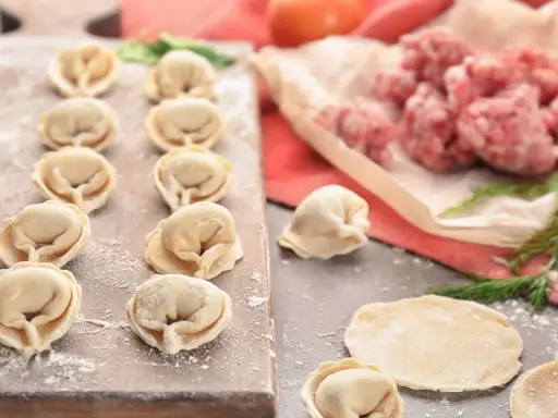 Gotowe do lepienia pierogi z mięsem mielonym surowym. Na desce ułożone pierożki, obok surowe mięso i ciasto.