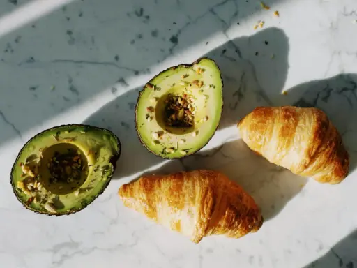 Dwa połówki awokado z nasionami i dwa chrupiące croissanty na marmurowym blacie. Idealne składniki na pyszne awokado przepisy.