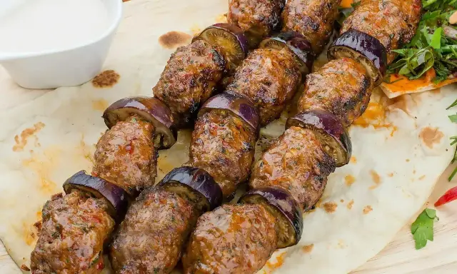 Kofta kebab z bakłażanem na patyku, podany na płaskim chlebie z sosem.