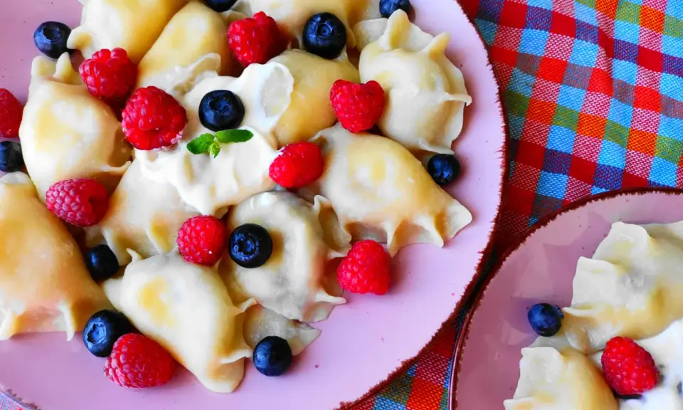 Pyszne pierogi z malinami i borówkami, polane śmietaną, na różowym talerzu. Letnia uczta pełna smaku.