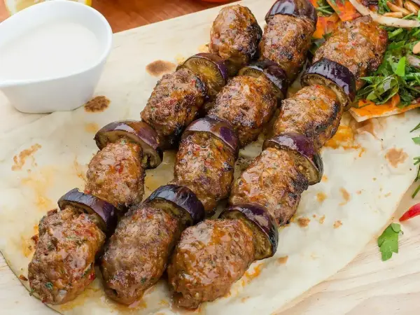 Kofta kebab z bakłażanem na patyku, podany na płaskim chlebie z sosem.